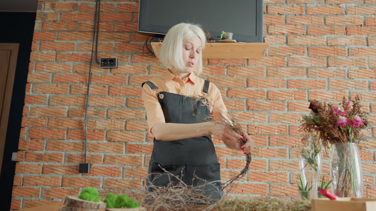 Woman in black apron preparing twigs for rustic floral craft arrangement on wooden table with moss and dried flowers, working carefully in cozy workshop with brick wall interior