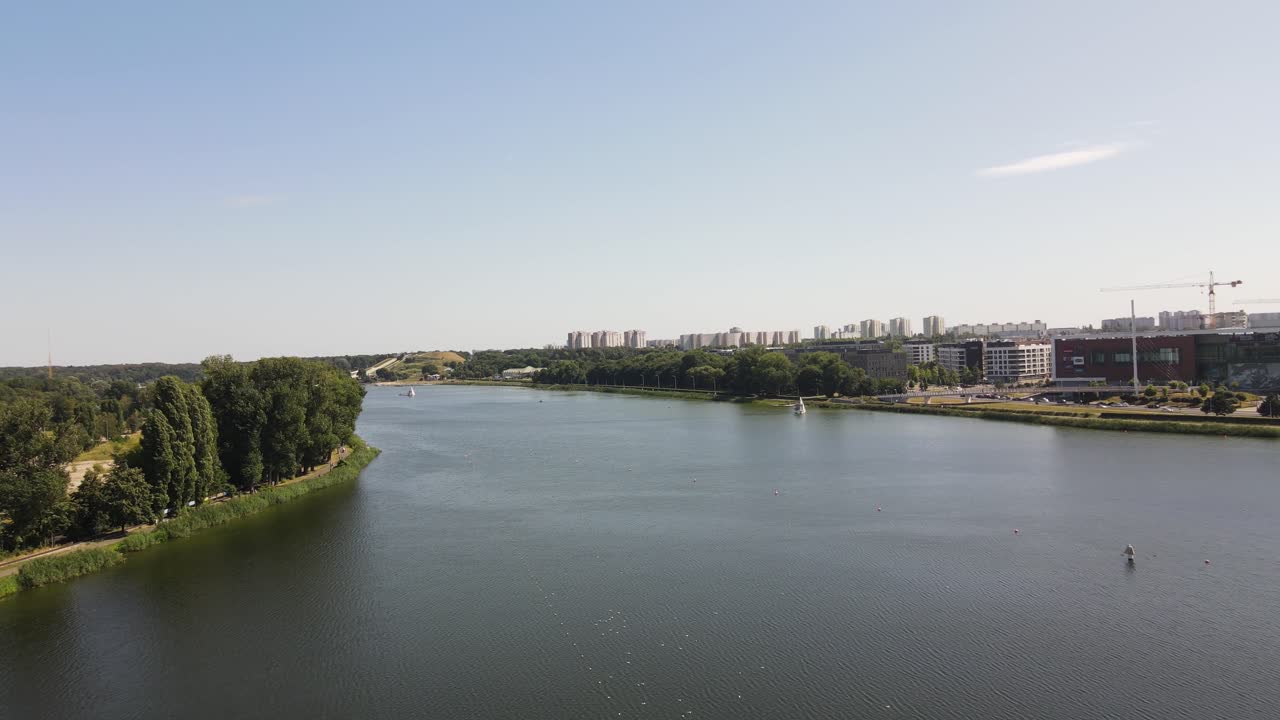 Video con drone del quartiere rivierasco di Varsavia, Polonia. Presenta acqua calma, skyline urbano, aree verdi e gru da costruzione sotto cieli sereni con riprese aeree stabili che catturano la scena.