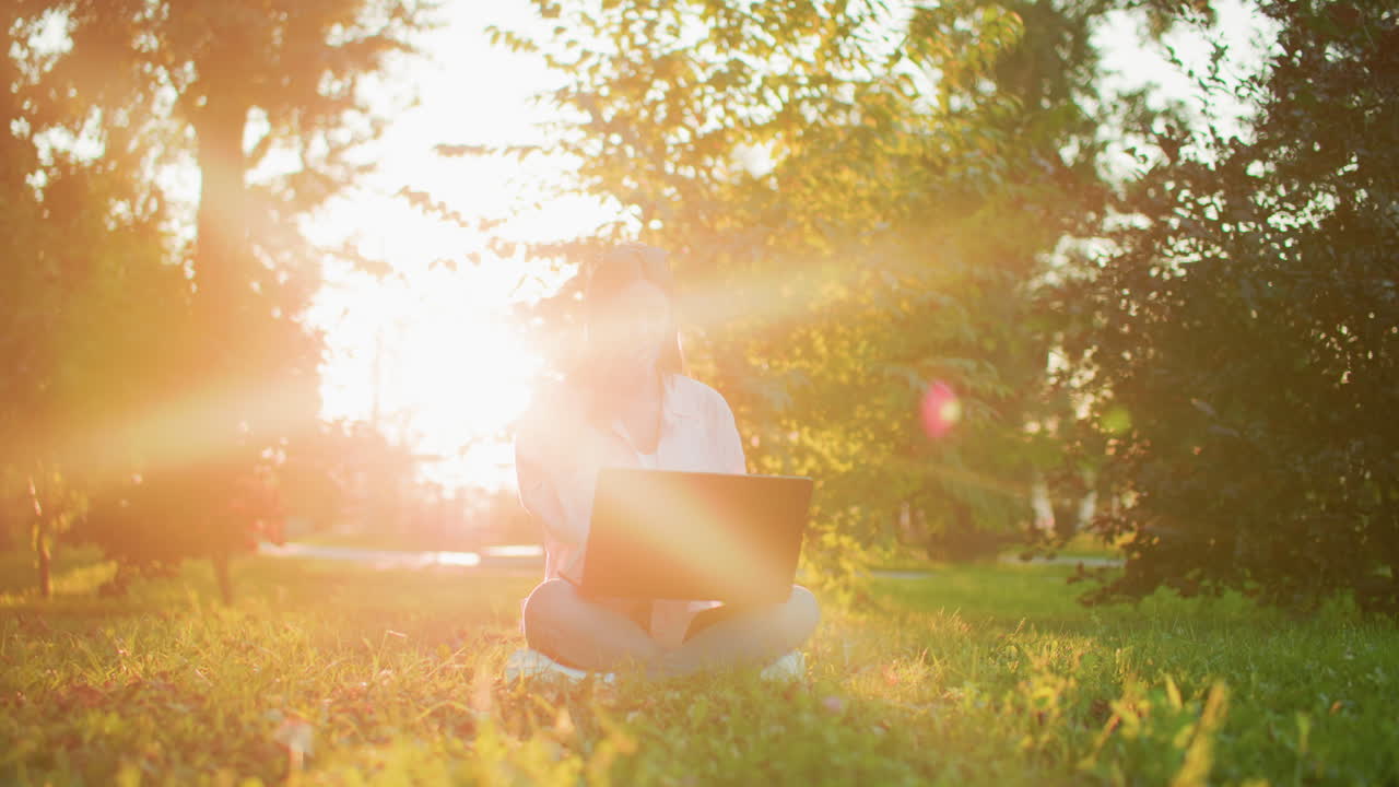 야외에서 노트북으로 일하는 프리랜서 여성, 빛이 나무를 통해 필터링하여 그녀의 주위에 황금빛을 만들어 잔디에 다리를 고 앉아 자연에서 평화로운 작업 환경을 즐기고 있습니다.