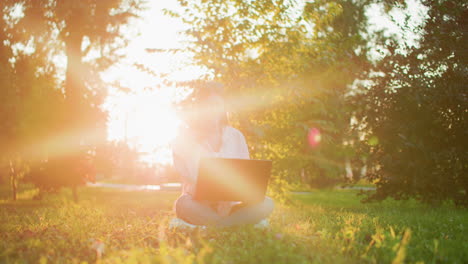 freelancer woman working on laptop outdoors with sunlight filtering through trees, creating golden glow around her as she sits cross-legged on grass, enjoying peaceful work environment in nature