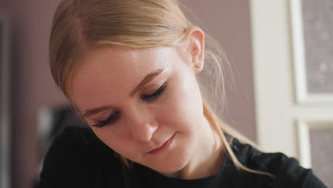 light skin blond-haired woman with middle-part hairstyle gently smiles while focusing on task, her hair tucked behind her ear, with soft blur of white panel door and pastel wall in background