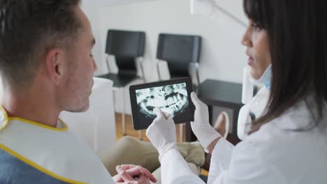 dentista biracial con máscara facial examinando los dientes de un paciente masculino en una clínica dental moderna