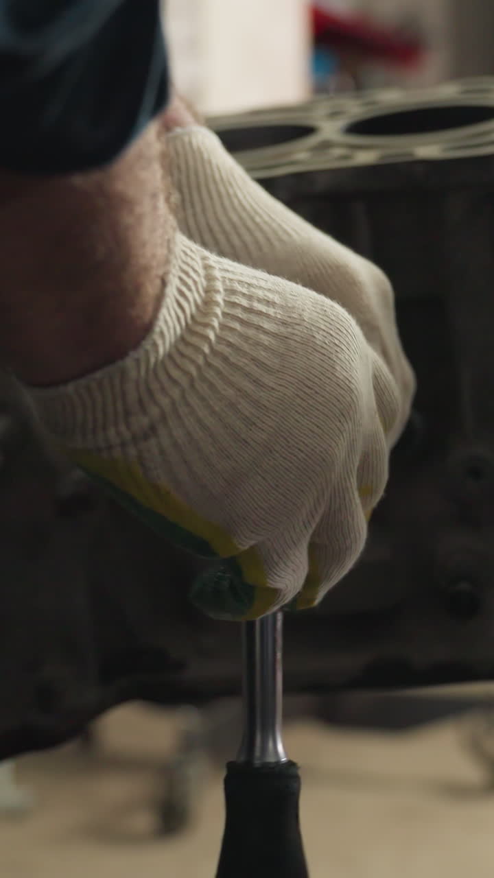 Close-up of mechanic in blue uniform using yellow scraper on car engine while another mechanic in white gloves loosens bolt, working together in busy automotive workshop