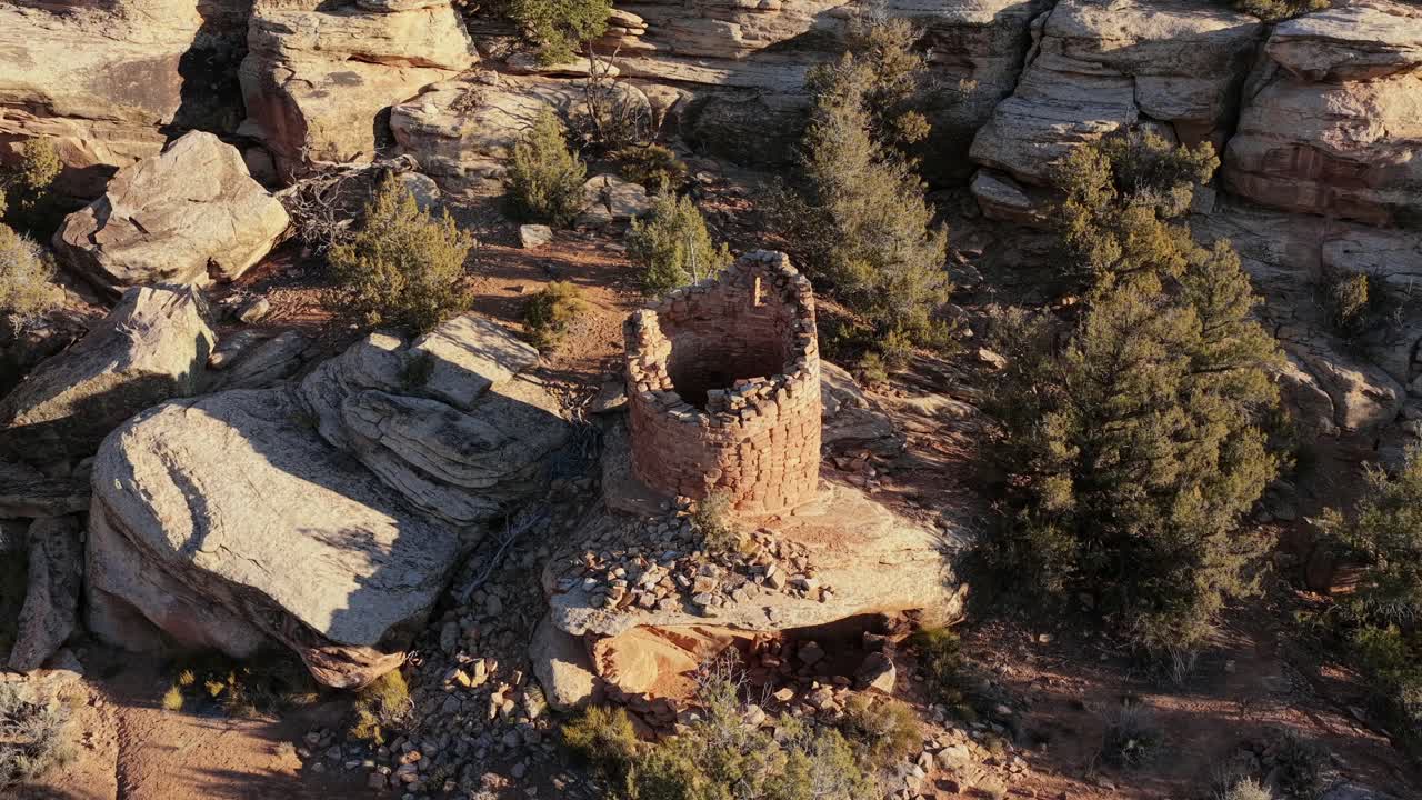 An ancient dry-stone tower sits perched on a rocky mesa above a deep desert canyon. The intricate masonry is surrounded by high-desert vegetation and rugged geological formations
