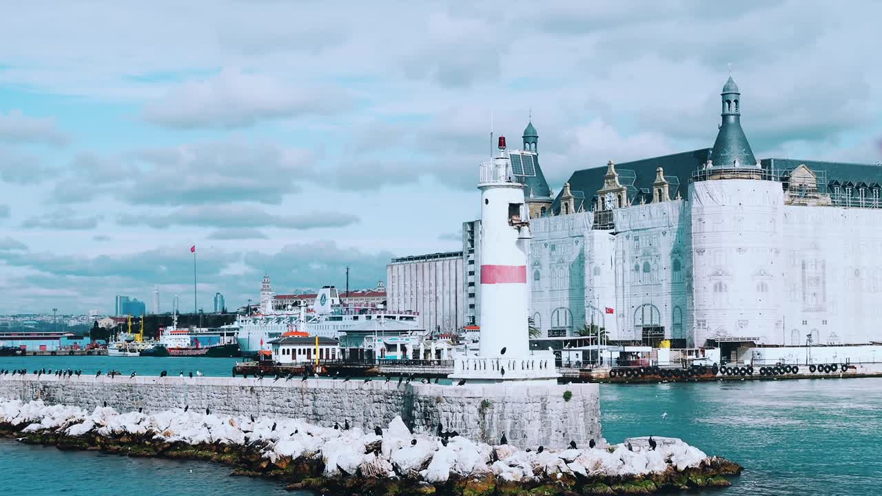Flight around modern white and red lighthouse in Istanbul harbour with flying seagulls. Mediterranean sea calm waves many concrete buoys pier breakwater navigation shipping port City landscape skyline