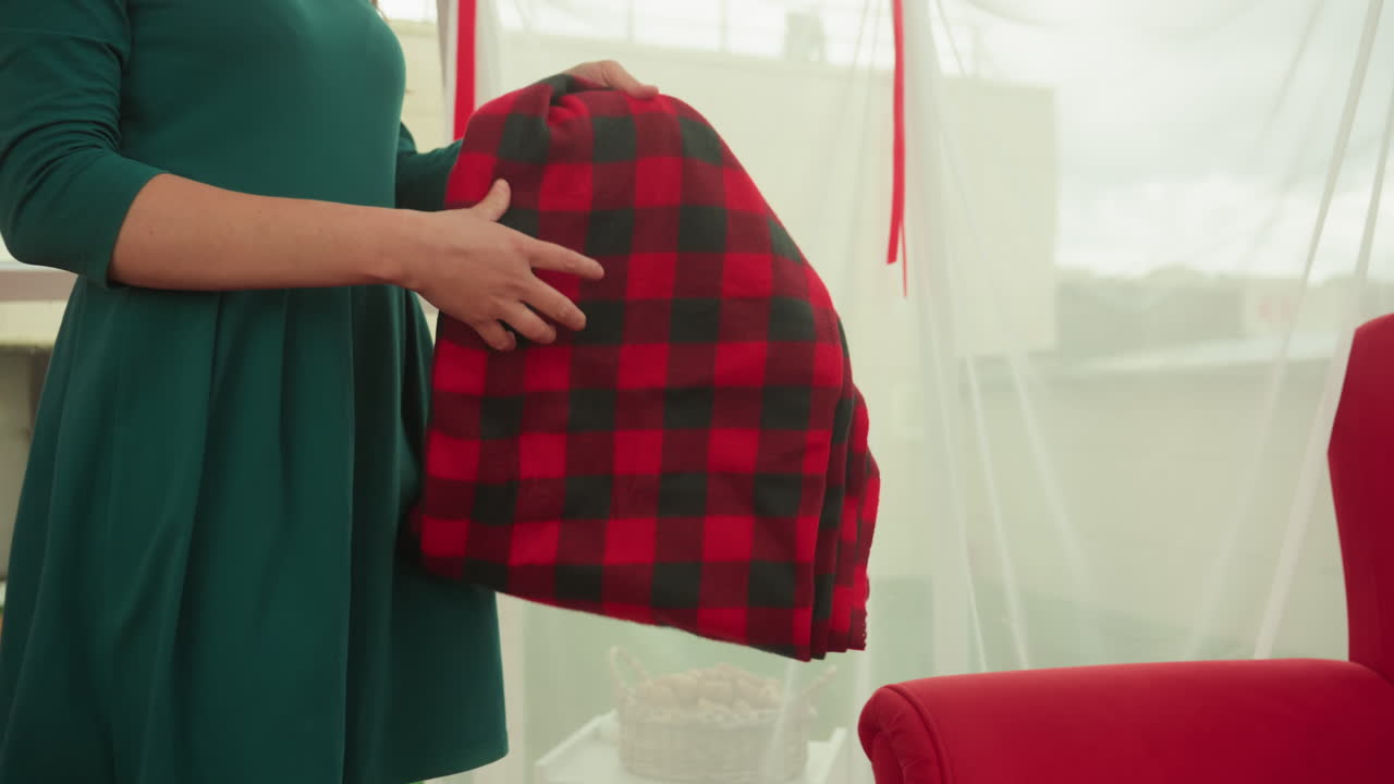 white woman brings blanket to sofa, emerald dress arranging plaid under canopy curtains in sunlit rooftop lounge, gentle movement and anxious anticipation, red upholstery and soft textiles create warm