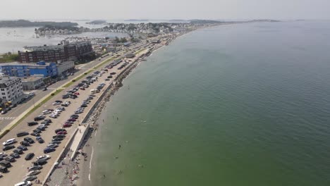 Pájaros-Volando-Sobre-La-Playa-De-Nantasket-En-Hull,-Massachusetts-Con-Gente-Nadando-Y-Autos-Estacionados-En-Verano