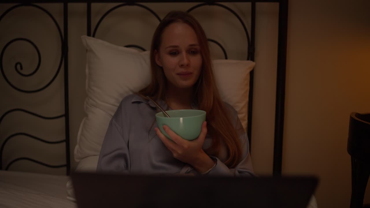 Donna che fa colazione a letto mentre guarda qualcosa sul laptop di notte