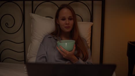 woman eating breakfast in bed while watching something on laptop at night