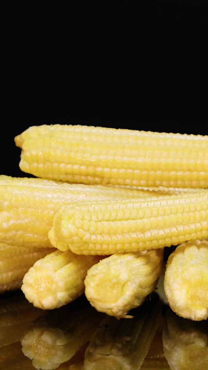 A pile of yellow baby corn rotates smoothly on a reflective black surface, illuminated by soft studio lighting, highlighting texture and freshness in close-up detail