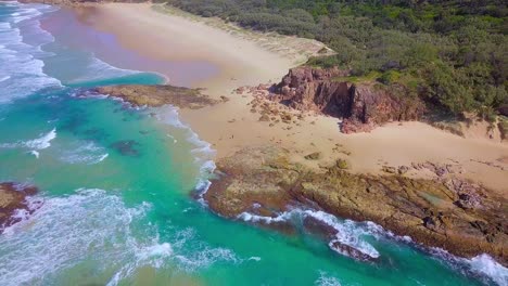 Aerial-fly-out-shot-of-a-wild-sandy-beach-with-cliffs,-rocks,-turquoise-blue-water-and-waves