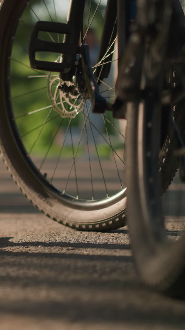 Close-up of lady's legs in pink sneakers kicking a bicycle tire to check air pressure, background showcases lush greenery and trees, casting shadows on the ground while a figure walks by