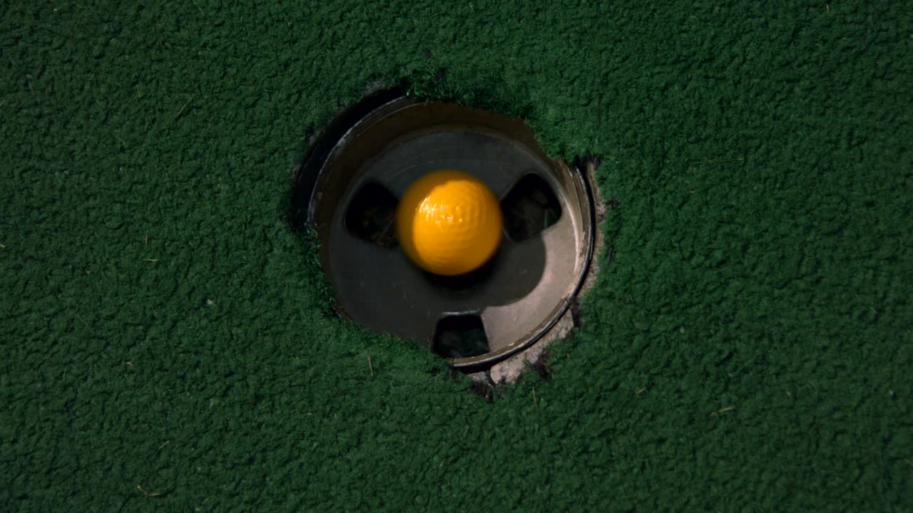 A close up birds eye view of a yellow mini golf ball falls into the golf hole and bounces in the pocket on a course