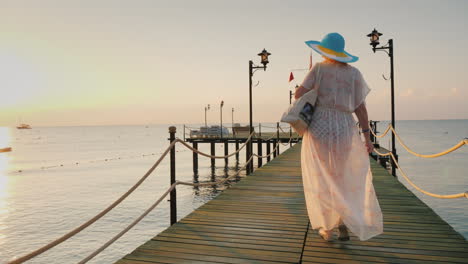 una mujer con un par y un sombrero camina por el muelle temprano en la mañana respira el aire fresco steadicam