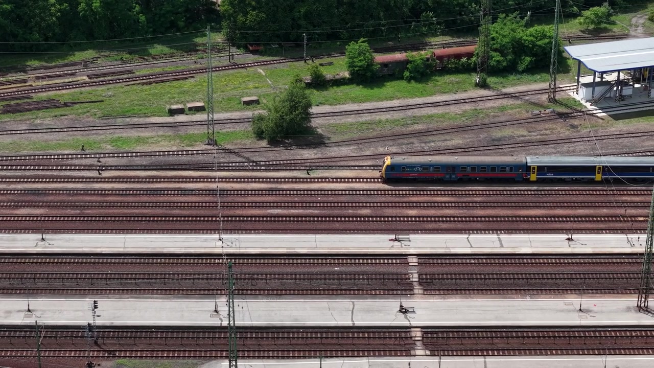 Una vista aerea dall'alto cattura un vasto snodo ferroviario con più binari, linee elettriche aeree e piattaforme nell'Ungheria rurale.