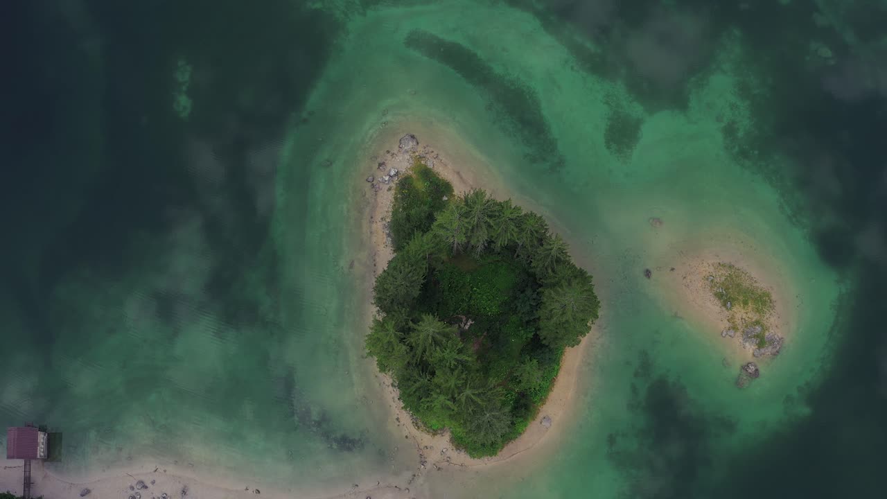 toma aérea de arriba hacia abajo de la isla en un lago alpino con una textura fantástica de agua verde esmeralda y nubes que se reflejan en la superficie