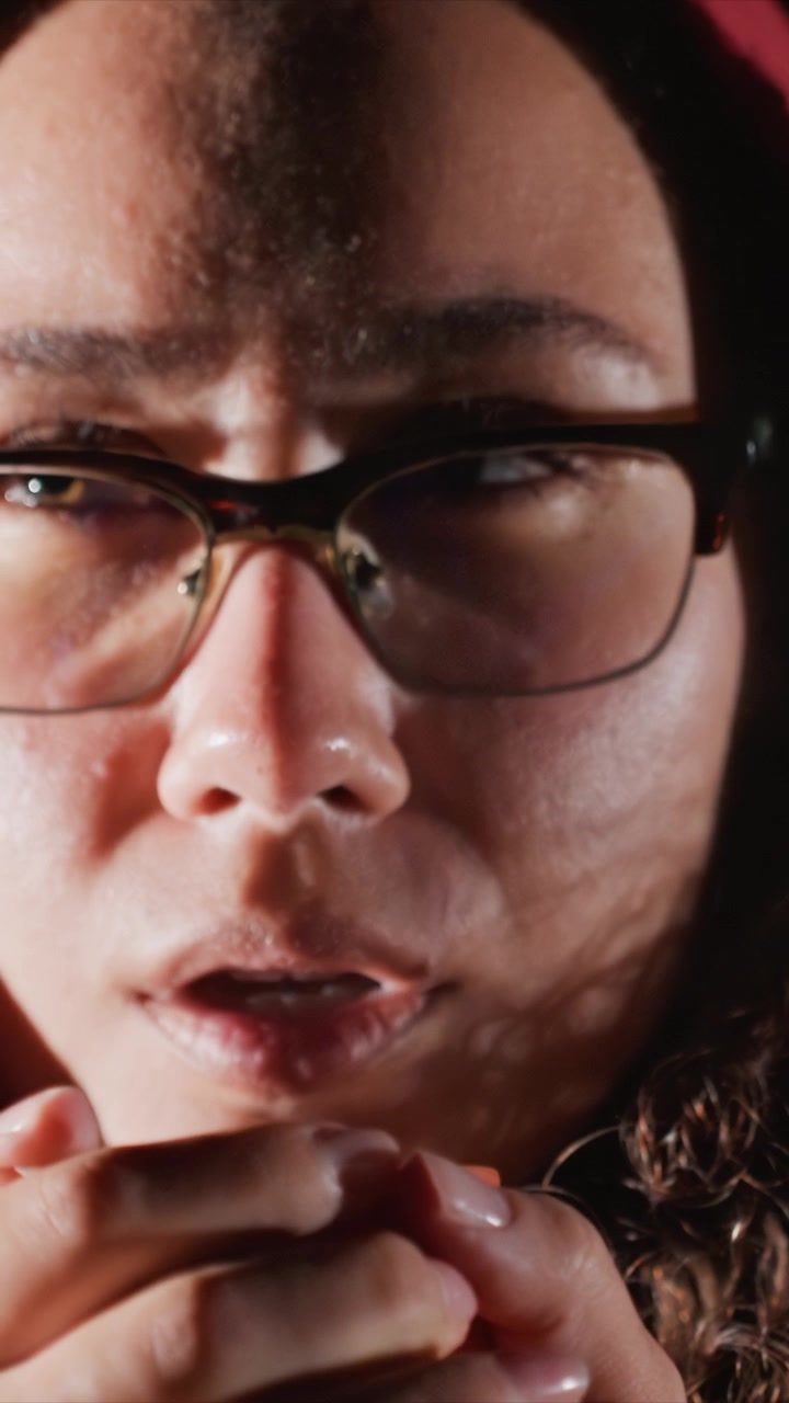 Person with visible eye lashes displays facial features. Glasses frames are dark nails on fingers appear. Shallow depth of field keeps attention solely on hands eyes nose