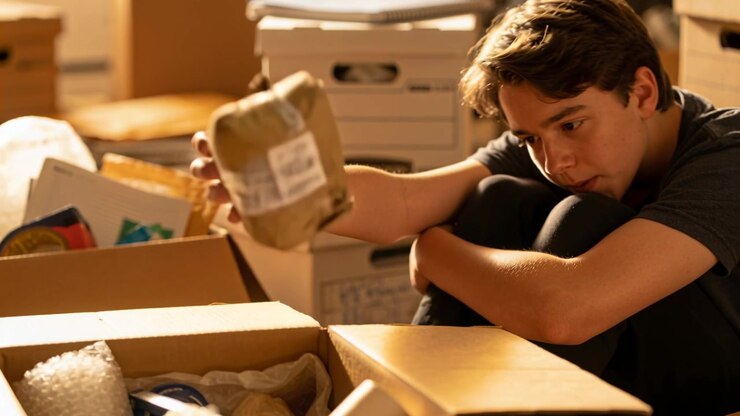 Seeing open moving box, teen in dark clothes reaching, inspecting wrapped bowl during home packing