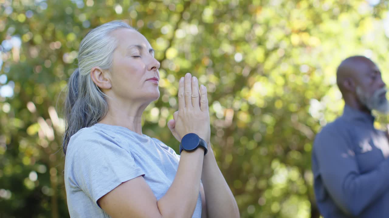 Ein Paar beginnt im Park mit der Meditation, wobei die Frau die Arme hebt und die Handflächen zur Brust senkt; eine runde Uhr ist zu sehen.