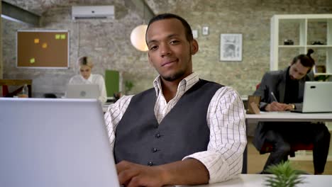jovem empresária africana está sentada no laptop no escritório, observando na câmera, colegas estão em rede com tecnologias, conceito de trabalho, conceito de comunicação
