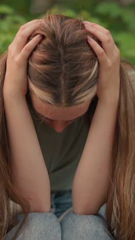 mujer sentada en un banco de madera se inclina hacia adelante, descansando la cabeza en sus manos, el cabello largo en cascada hacia abajo, parece reflexiva e introspectiva, con vegetación y árboles en el fondo