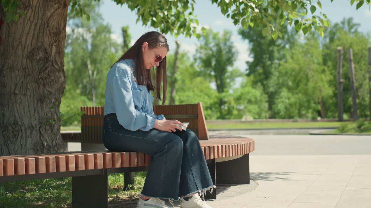 백인 여성 학생이 밖에서 레슨을 계획하고 있어요. 백인 여성이 나무 옆에 앉아 교과서를 공부하고 있고, 백인 여성이 나무 아래 굽은 벤치에 앉아 야외에서 교육 활동을 신중하게 계획하고 있어요.