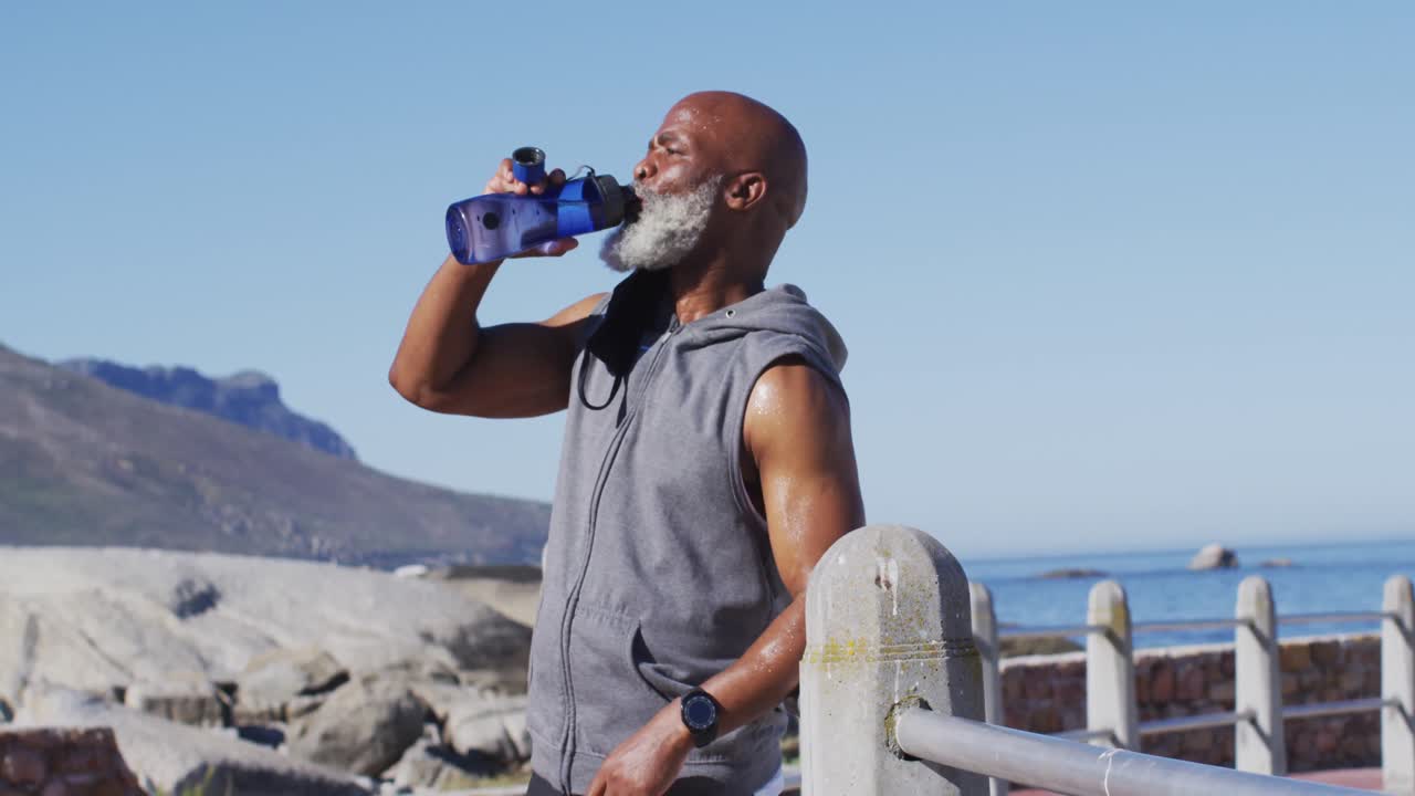 hombre afroamericano mayor ejercitando agua potable en rocas junto al mar