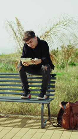 man picking up notes from the backpack and starting to study on the park bench