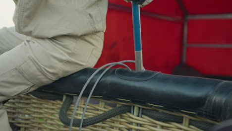 Close up of seated technician pushing metal stick into balloon basket fitting while cables loop across wicker frame, working carefully beside red truck in open rural area under soft natural daylight