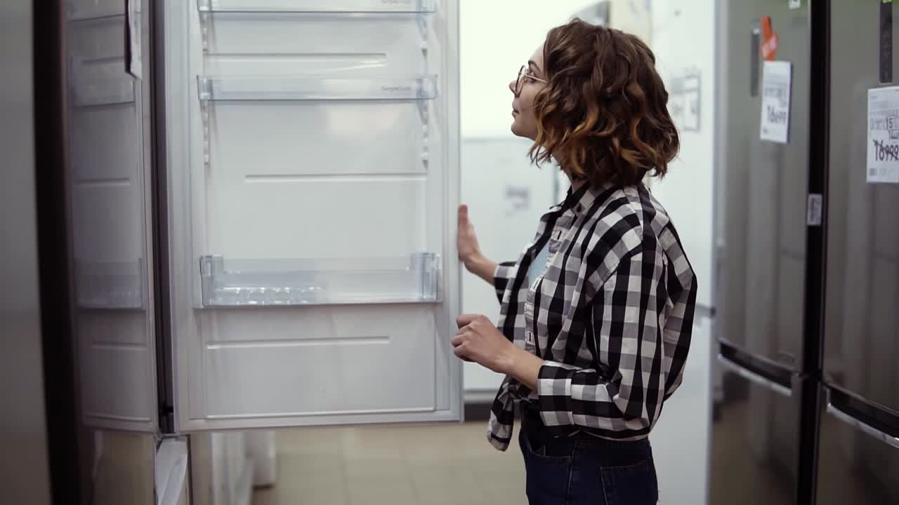 jovencita alegre seleccionando refrigerador doméstico en el supermercado. mujer rizada en camisa a cuadros decidiendo comprar uno nuevo para