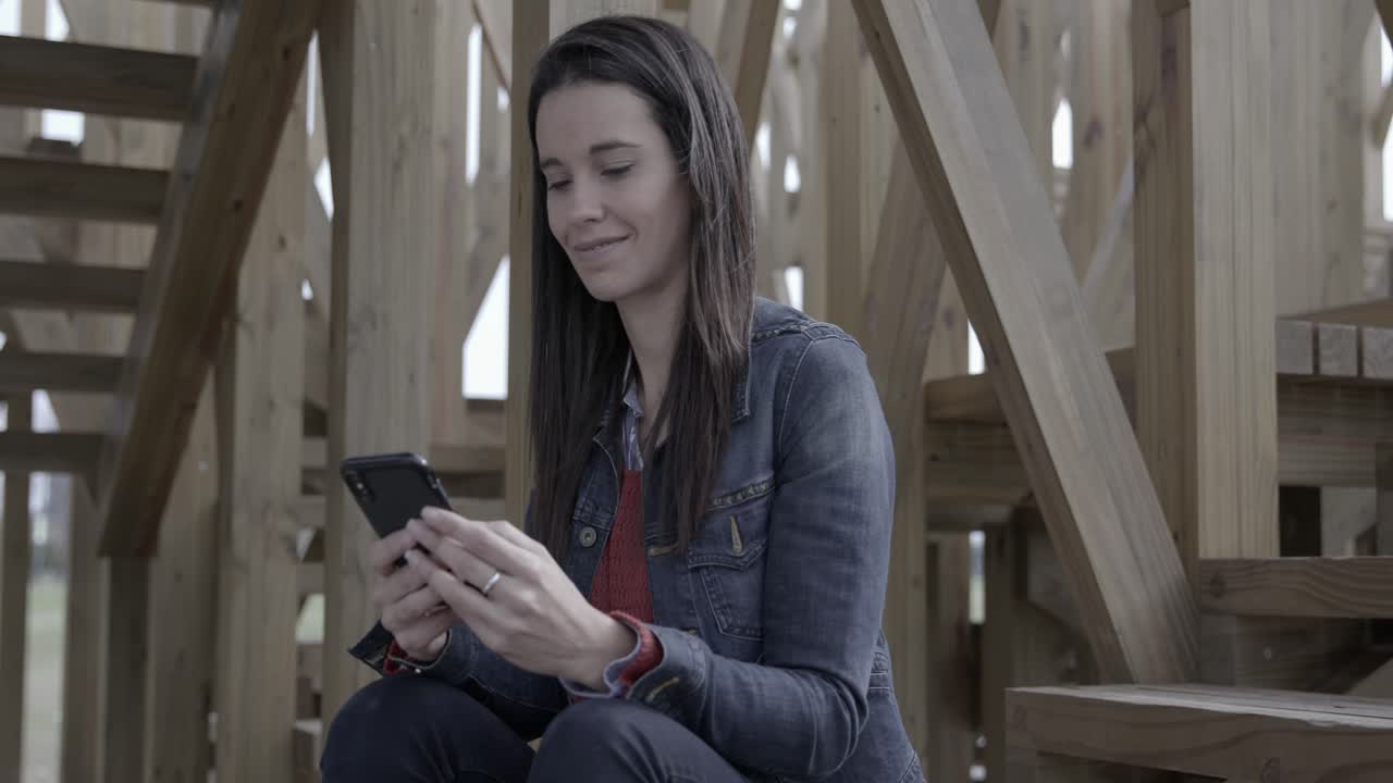 Joven sonriendo mientras usa un smartphone al aire libre bajo una estructura de madera