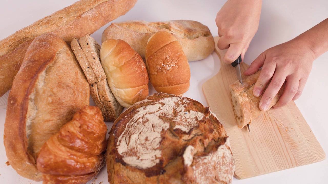 Hands slice crusty baguette on wooden board surrounded by sourdough, croissants, and artisan rolls.