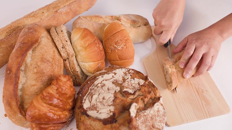 Hands slice crusty baguette on wooden board surrounded by sourdough, croissants, and artisan rolls.