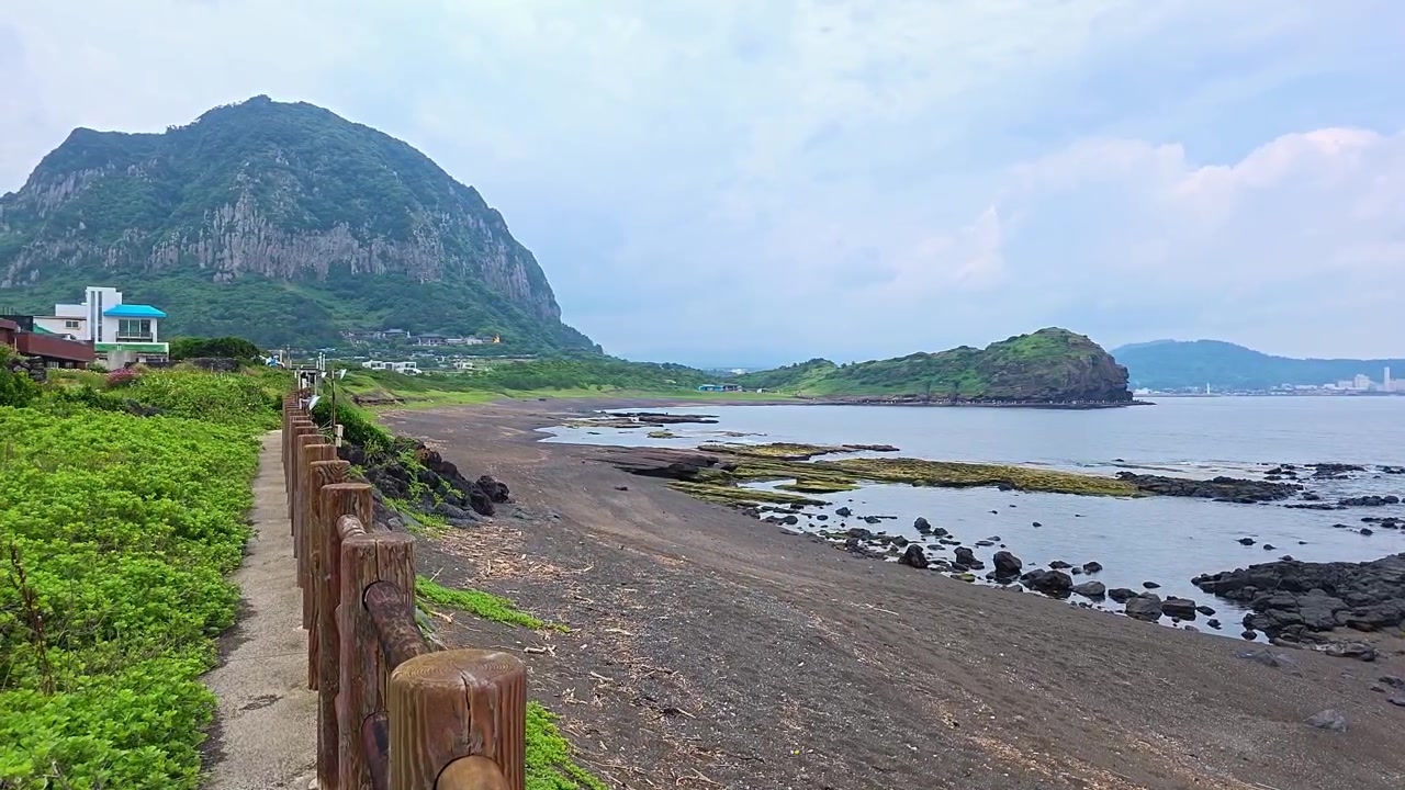 Prospettiva in prima persona mentre si cammina lungo il sentiero costiero della spiaggia di Sagye, con una recinzione di legno, oltre rocce vulcaniche, verso il monte Sanbangsan e il promontorio di Yongmeori a Jeju, Corea del Sud.