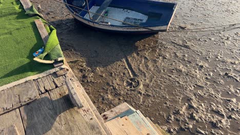 escena de caminar por un viejo y deteriorado paseo marítimo de madera con superficie de suelo seca y fangosa, viejo barco de pesca en el fondo vacío del río, captura diurna de estructuras desgastadas por el tiempo