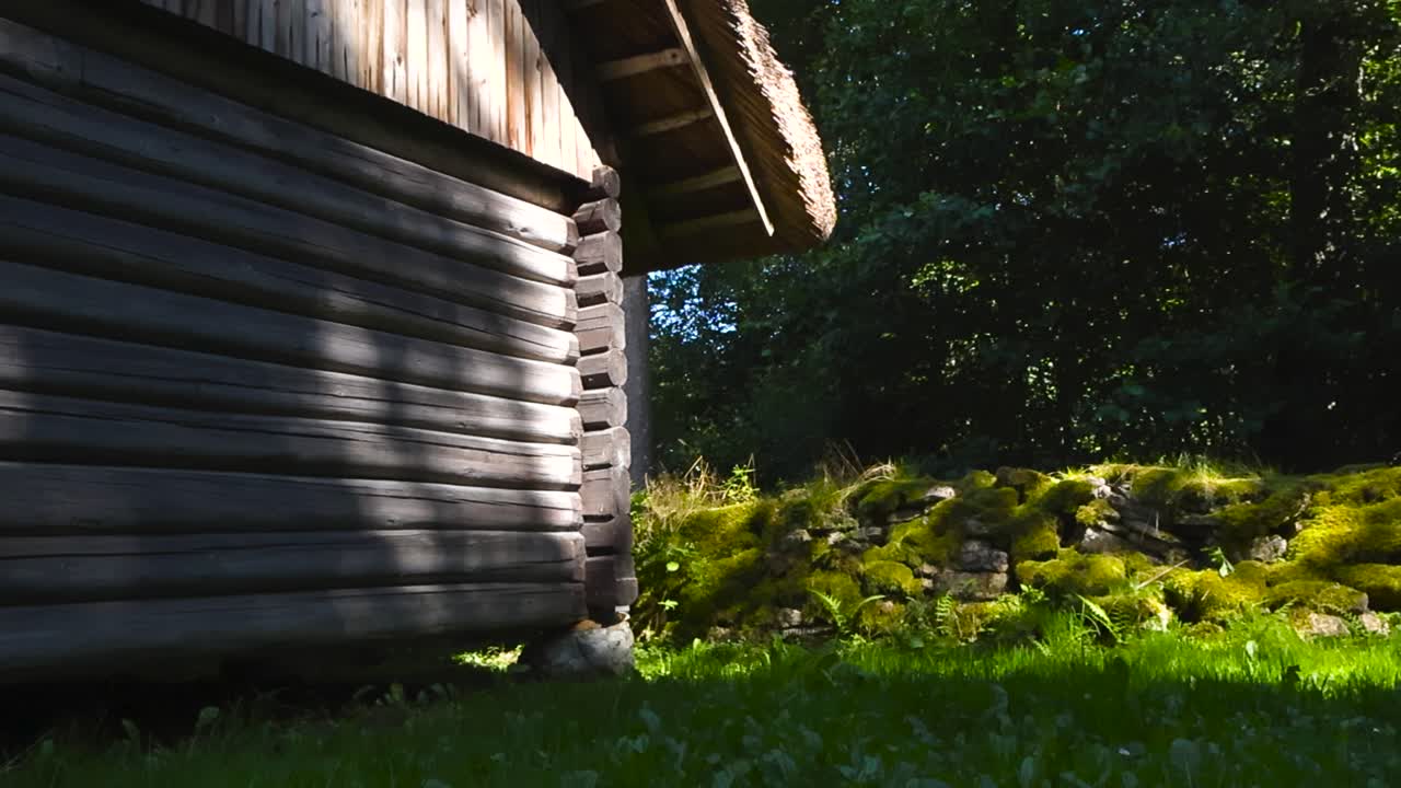 Low angle slow pan of traditional log building with thatch roofing and green moss covered dry stone wall at the Estonian Open Air Museum. Rocks holding up old farm house cottage, summer grass backyard
