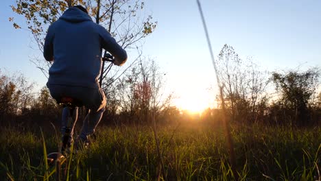 man riding a electric scooter over fields, in forest, during sunset - low, dolly view