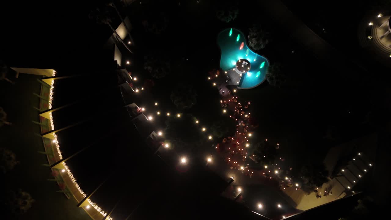 Aerial view of a bride and groom cutting the cake at night by a lit-up lake, surrounded by guests holding sparklers
