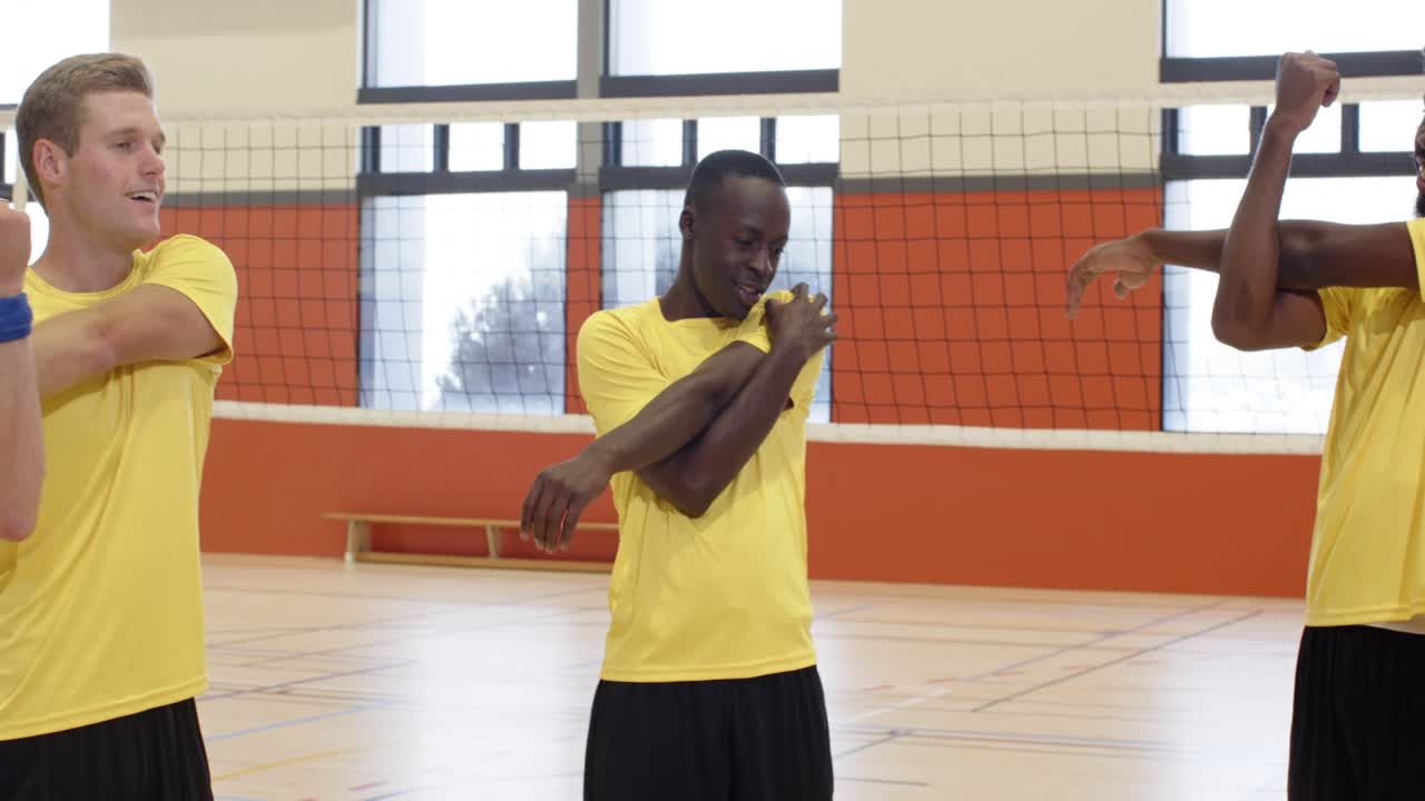 Diverse men in yellow-shirts copying blue-band leader doing crossbody stretch in gym by net warming