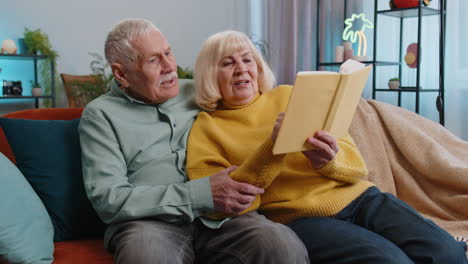 Senior-family-couple-man-woman-enjoying-reading-interesting-book,-talking,-laughing-at-home-on-sofa