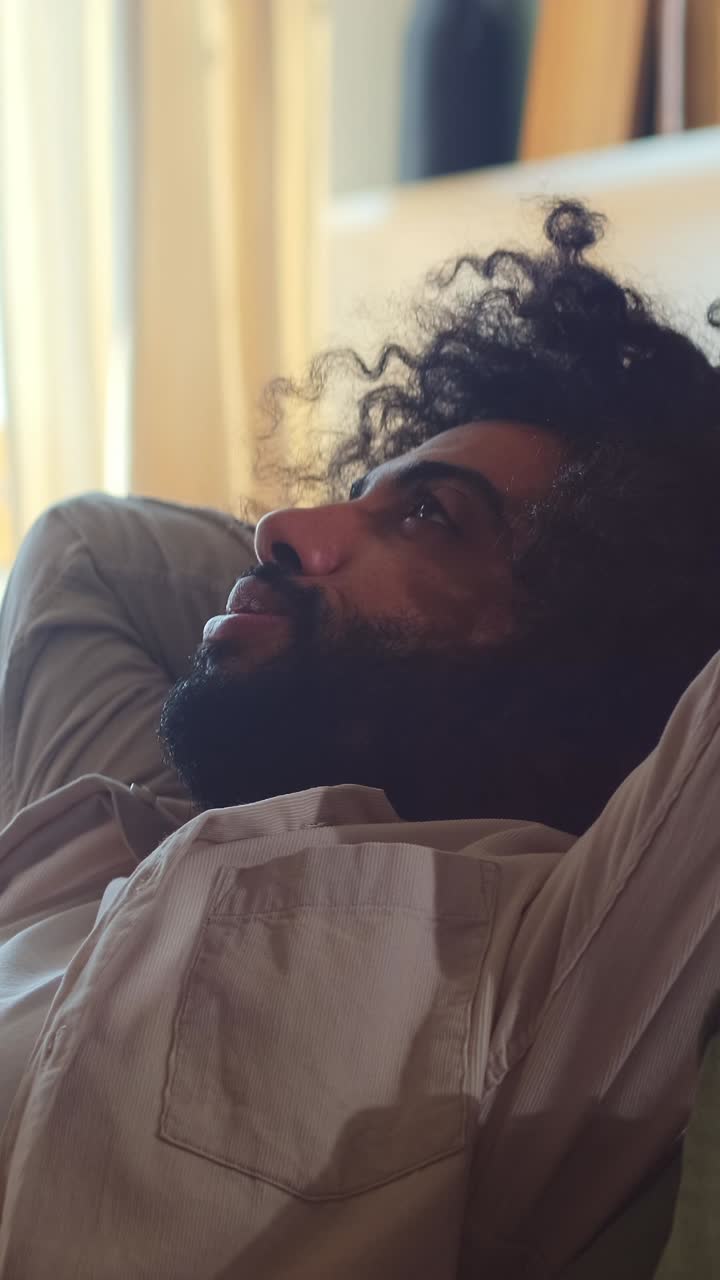 Ung, træt, positiv arabisk mand læner sig tilbage på sofaen for at slappe af efter en trættende dag.