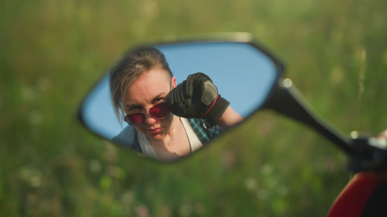 un reflejo en el espejo de una motocicleta que muestra a una mujer con una cálida sonrisa ajustando sus gafas de sol, está en un campo cubierto de hierba