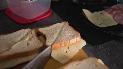 caregiver slices cheese sandwich with sharp knife on wooden board in kitchen, preparing meal with visible sliced bread, cheese, deli meat, and containers nearby, demonstrating homemade food preparation process