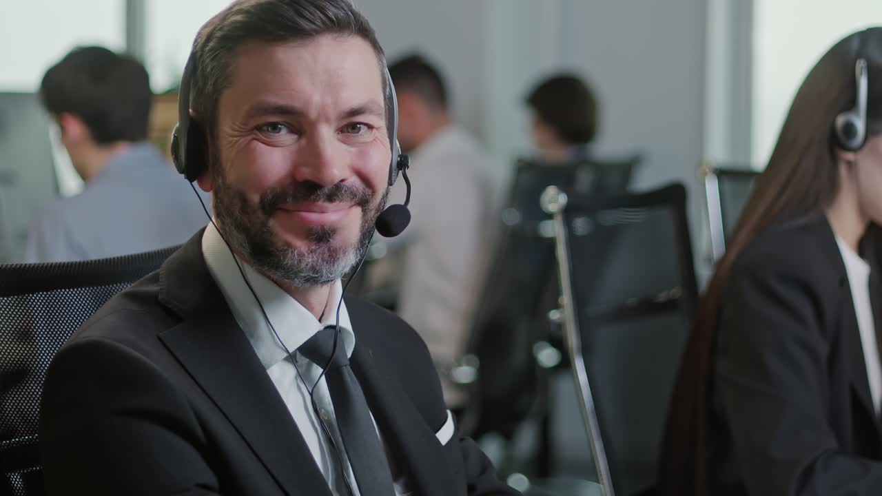 Portrait of Handsome Man Operator Look at Camera Smie Taking Calls Smile in Call Center Office. Use desktop pc at computer table and phone line at modern co-working