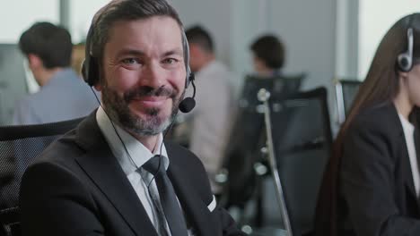 portrait of handsome man operator look at camera smie taking calls smile in call center office. use desktop pc at computer table and phone line at modern co-working