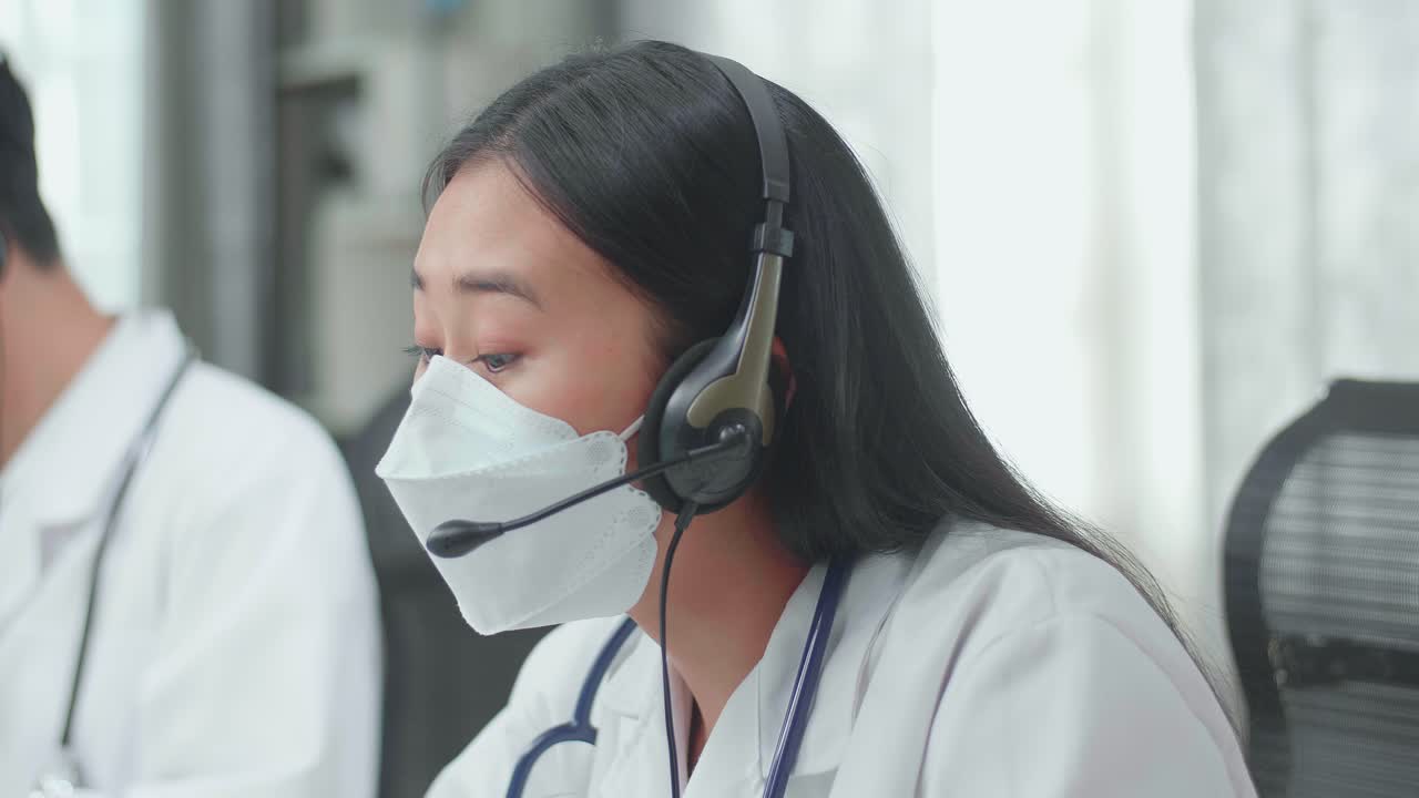 Close Up Of An Asian Woman Doctor In Headsets And Masks Working As Call Center Agent Speaking To Customer On The Call During Woking With Her Colleagues At The Office
