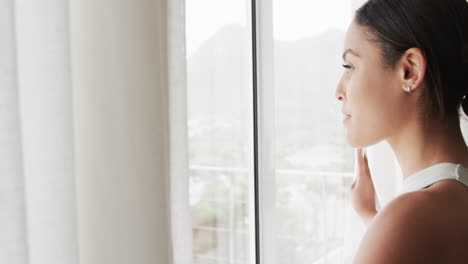 Happy-biracial-woman-looking-through-window-at-home,-slow-motion