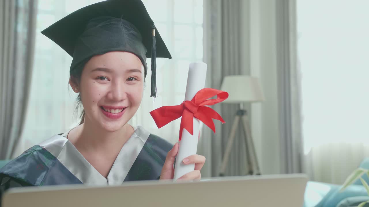 Excited Asian Woman Smiling And Showing Off A University Certificate To Camera. Pretty Female Graduate Wearing A Graduation Gown And Cap Sitting On The Living Room