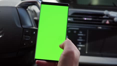 mano cercana del conductor en el coche utiliza un teléfono inteligente con una pantalla verde, dentro de un coche.