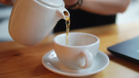 Cierra-La-Mano-Femenina-Sirviendo-Té-Caliente-De-La-Tetera-En-La-Taza-Blanca.-La-Cámara-Se-Mueve-Y-Muestra-A-Una-Hermosa-Joven-Con